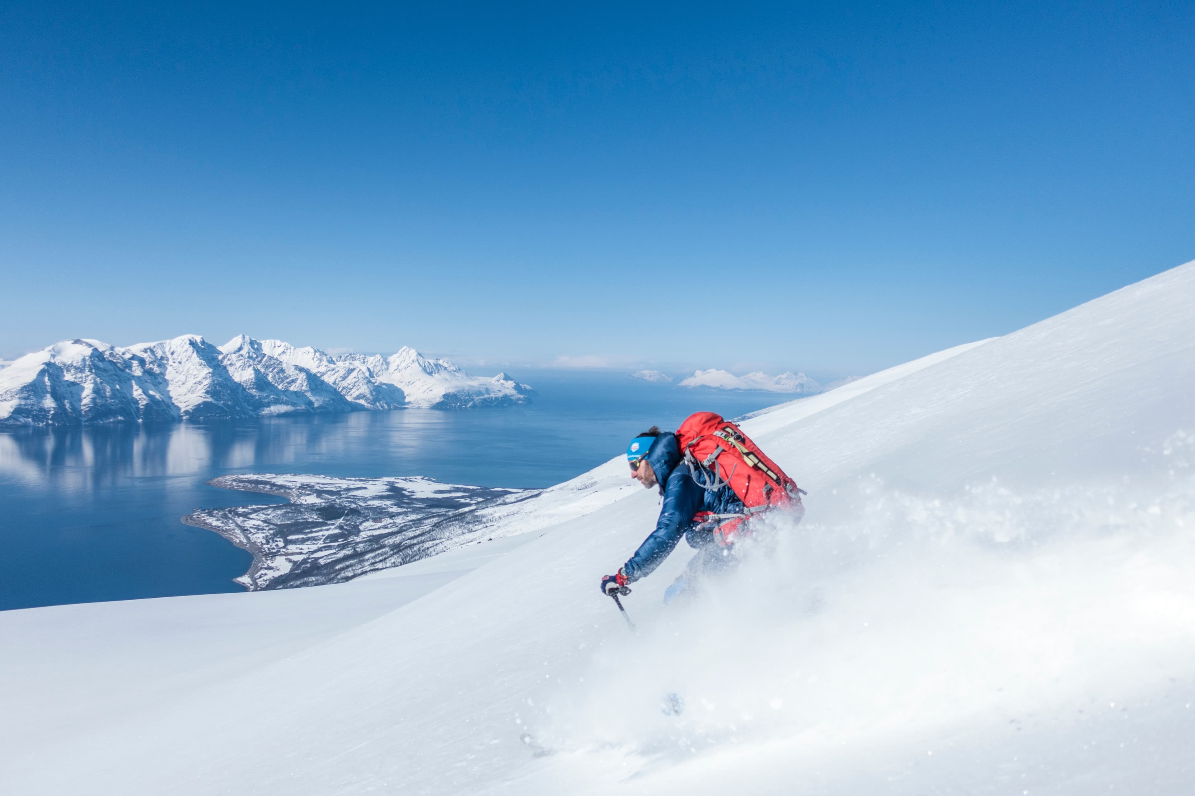 Een skiër met een rode rugzak daalt een besneeuwde berghelling af onder een strakblauwe hemel, met een prachtig uitzicht op besneeuwde bergen en een kalme blauwe zee op de achtergrond.