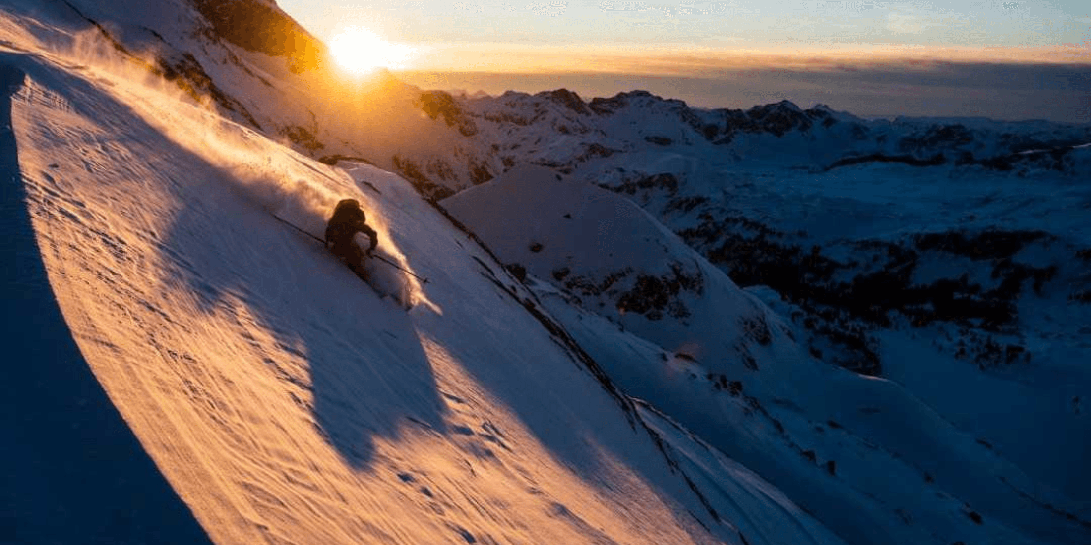 Trois paires de skis sont soigneusement disposées sur une terrasse en bois, accompagnées de divers équipements de ski. De gauche à droite : une paire de skis bleus, une pelle à neige rouge, une paire de skis verts, des bâtons de ski, un détecteur d'avalanche et une paire de skis noirs et blancs. Sous les skis se trouvent des paires de chaussures de ski rouge/noir et argent/vert.